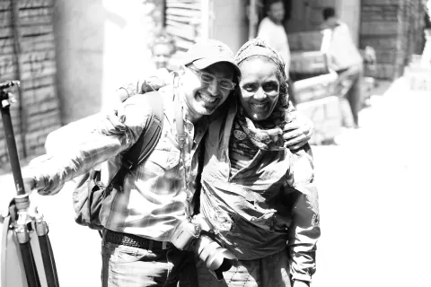 Two smiling men with equipment on a street, photographed in black and white.