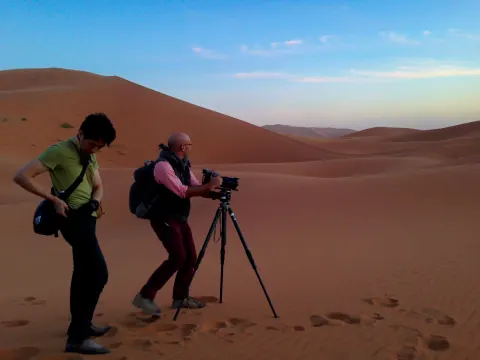 A photographer leaning over a tripod camera on a desert dune.