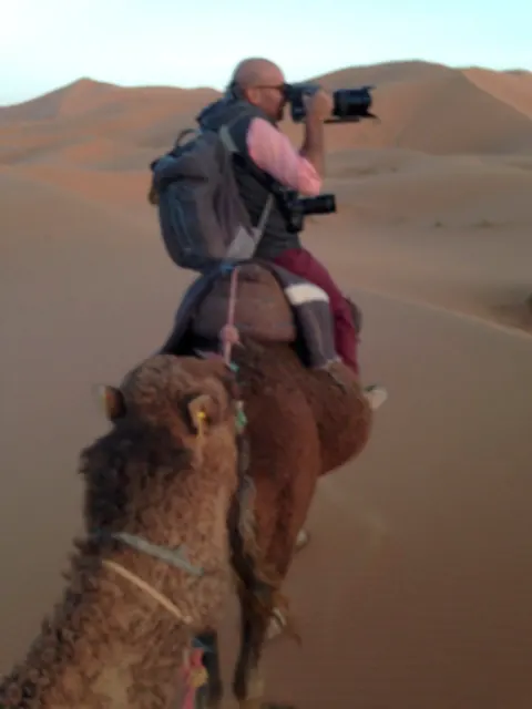 A photographer riding a camel while holding a camera.