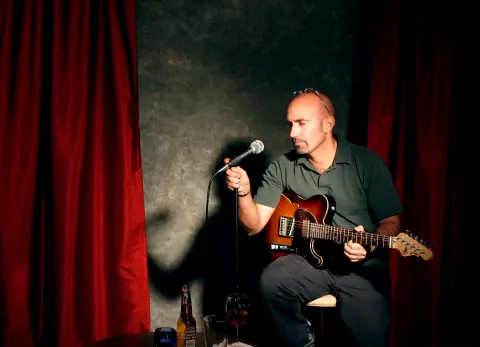A seated man holding a guitar and microphone beside red curtains.