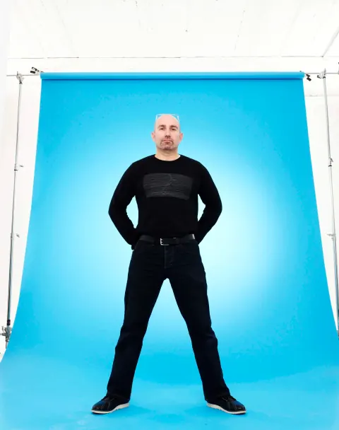 A man dressed in black posing against a blue studio backdrop.