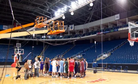 A group gathered beneath raised lighting equipment in a basketball arena.
