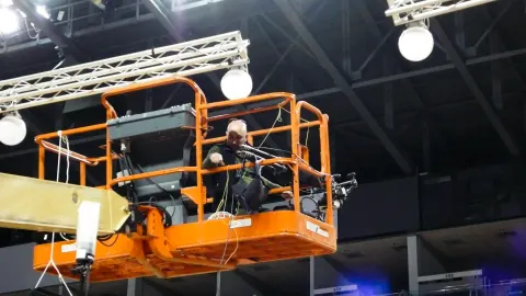 A person in an orange elevated work platform beneath arena lighting.