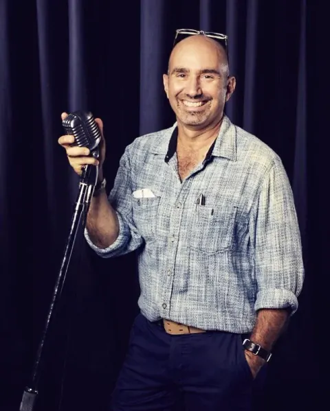 A smiling man holding a camera in front of a dark curtain.