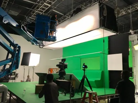 A camera crew filming in a studio with a green screen and overhead diffusion.
