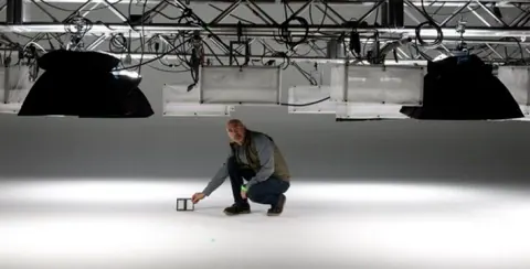 A man crouching on a white studio floor under suspended lights.