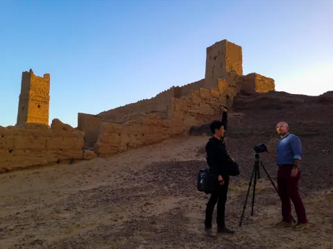 Two people beside a tripod at the foot of a fortification wall.