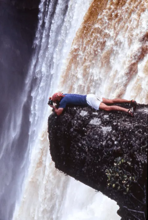 A person lying on a projecting rock beside a waterfall.