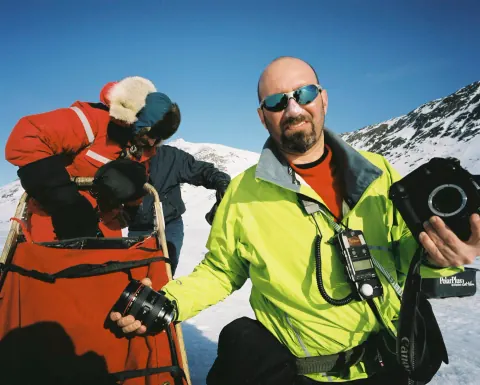 A smiling photographer in a bright jacket beside snowy mountains.