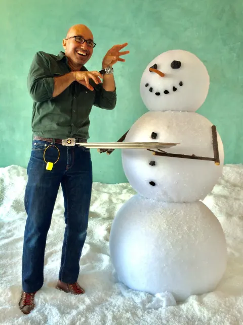 A man gesturing beside a large snowman on a studio set.