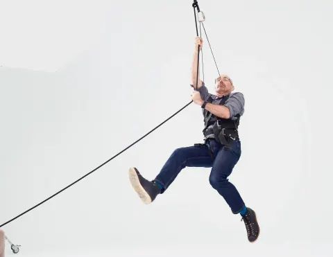 A man suspended from ropes against a white studio backdrop.