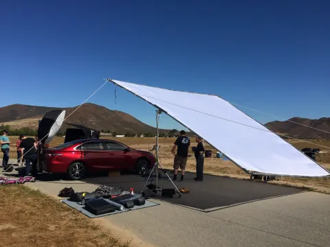 A large white reflector beside a red car at an outdoor shoot.