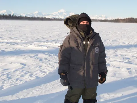 A bundled-up person standing in a snowy field with mountains in the distance.