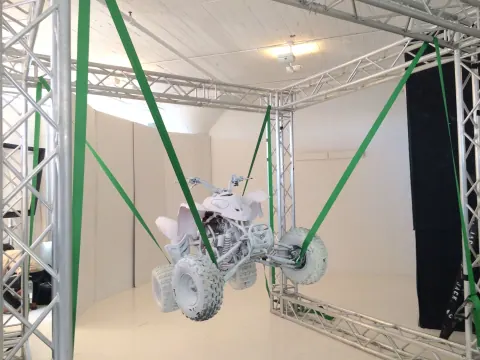 A small vehicle suspended by green straps inside a studio rig.