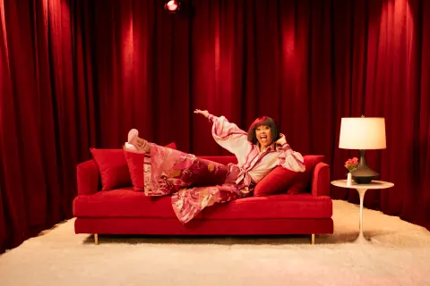 A woman in pink reclining on a red sofa in front of red curtains.