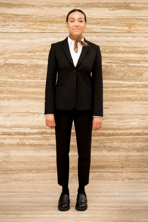 A woman in a black suit and white shirt standing against a pale stone wall.
