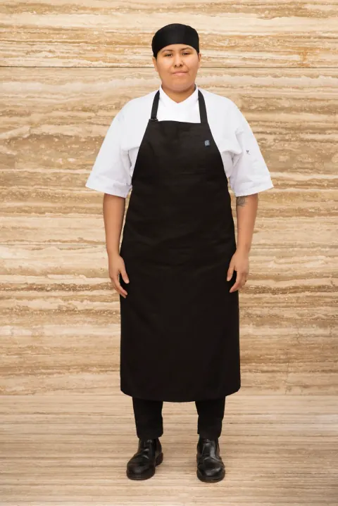 A person in a black apron and white shirt standing against a pale stone wall.