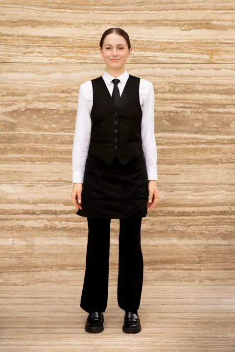 A woman in a black waistcoat and tie standing against a pale stone wall.