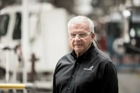 A grey-haired man in a black polo shirt on a factory floor.