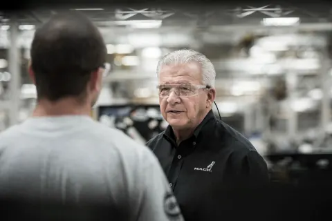 A grey-haired man talking with another person among factory machinery.