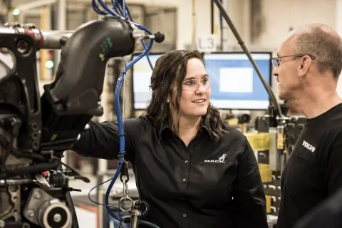 A woman and man in black polo shirts talking beside factory equipment.