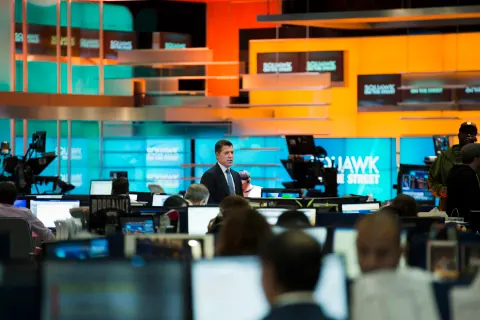 A broadcast studio with a presenter at a desk and orange and blue screens.