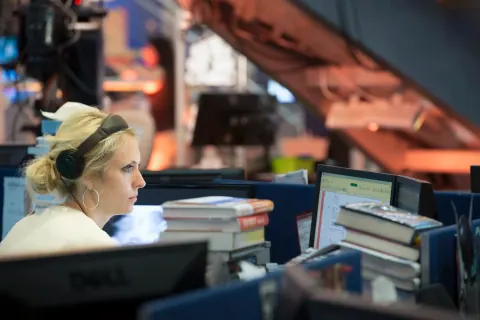 A woman working at a desk with several monitors in a newsroom.