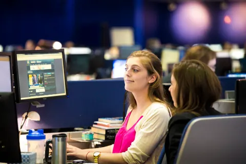 A woman in a bright pink top and pale cardigan at a newsroom desk.