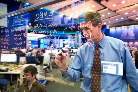 A man writing on a glass board in a newsroom.