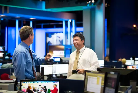 Two men talking among desks and screens in a broadcast newsroom.