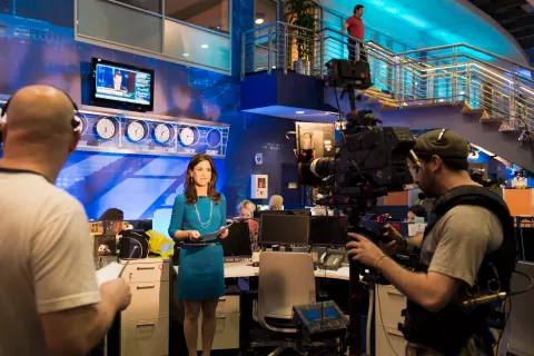 A presenter in a teal dress being filmed by a television camera crew.