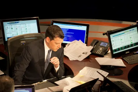 A man in a suit reviewing papers at a desk surrounded by monitors.