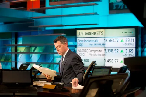 A suited presenter at a broadcast desk in front of market screens.