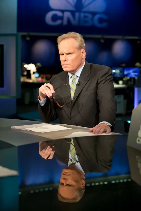 A suited man at a reflective broadcast desk beneath a CNBC sign.