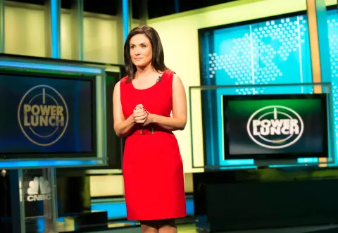 A woman in a red dress in front of Power Lunch studio screens.