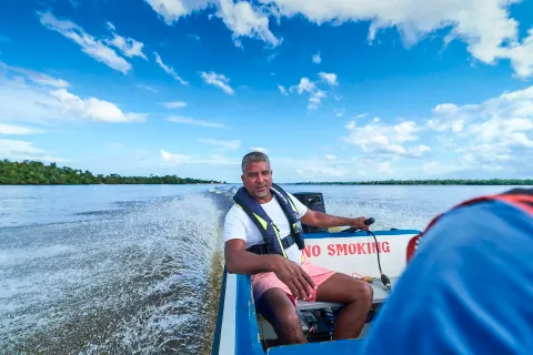 A man sitting in a moving boat as its wake crosses a wide river.