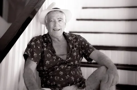 A black-and-white portrait of a man in a hat seated beside a staircase.