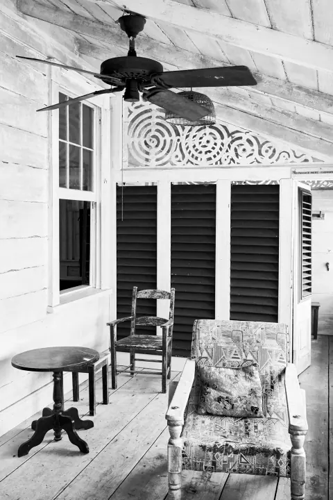 A monochrome veranda with shutters, a ceiling fan and a wicker chair.