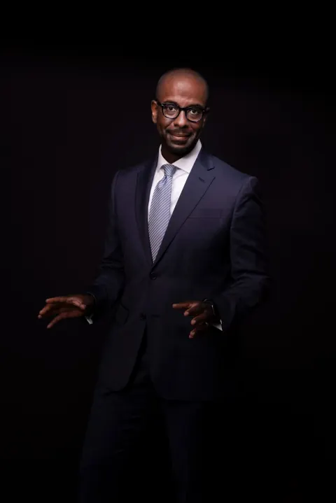 A seated man in a dark suit and glasses against a black background.