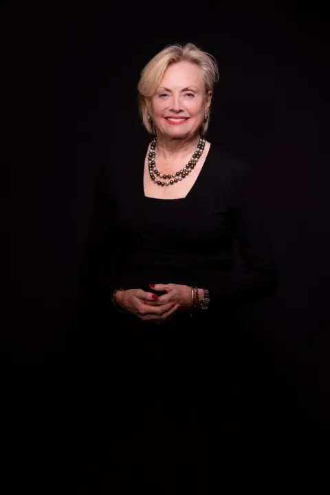 A smiling woman in a black outfit and necklace against a dark background.