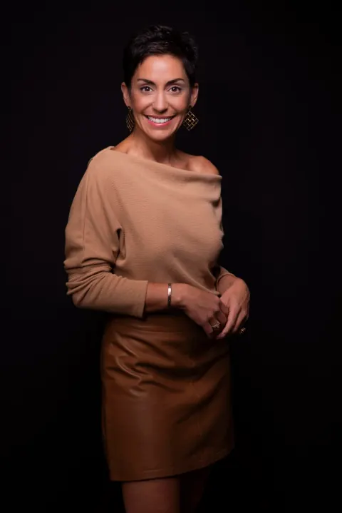 A woman in a tan top and brown skirt with her hands clasped.