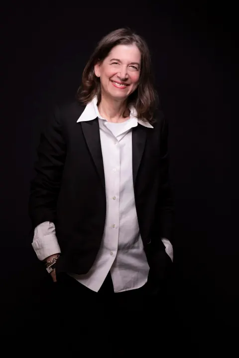A smiling woman in a black blazer and white shirt, with her hands in her pockets.