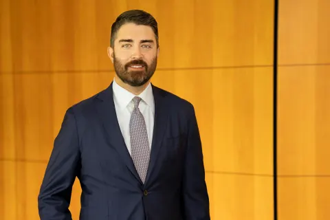 A bearded man in a navy suit and tie against a wood-panelled wall.