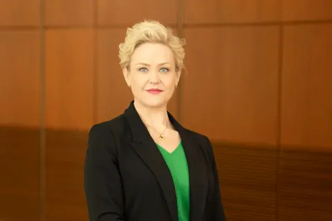 A woman in a black blazer and green top against a wood-panelled wall.