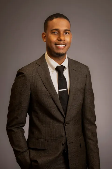 A smiling man in a dark suit and black tie with one hand in his pocket.