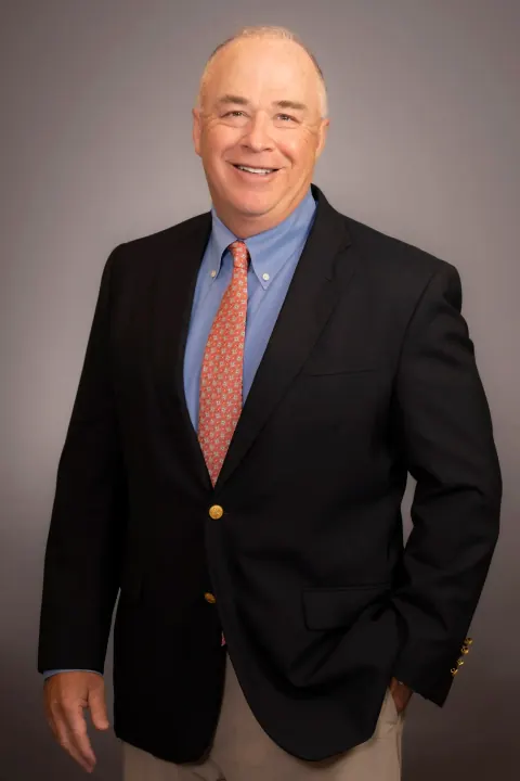 A smiling man in a dark blazer, blue shirt and orange tie.