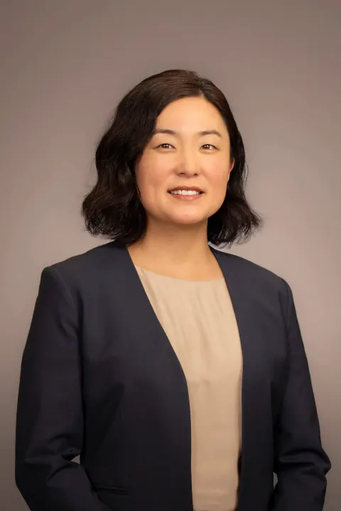 A woman in a navy blazer and beige blouse against a grey backdrop.