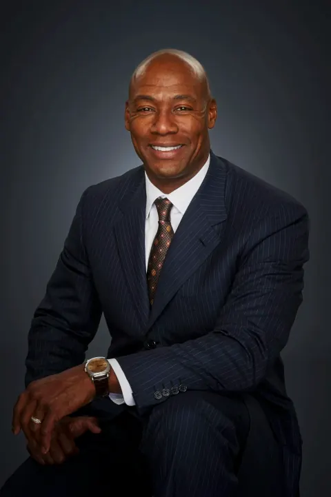 A smiling seated man in a navy suit with his hands resting on his knee.