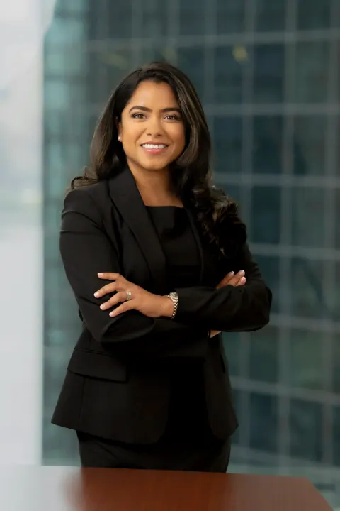 A smiling woman in a black blazer with folded arms beside office windows.