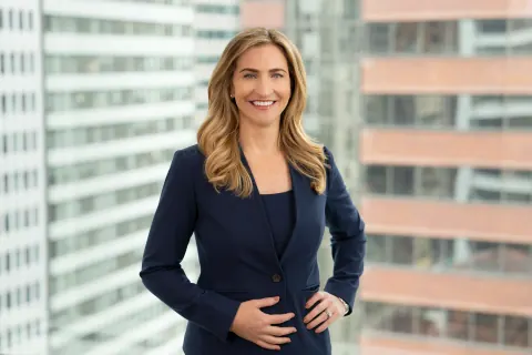A smiling woman in a navy suit in front of city-facing office windows.